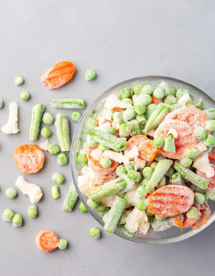 Frozen vegetables in a glass bowl and on a table, green peas, carrot, cauliflower,  green beans, top view, square format